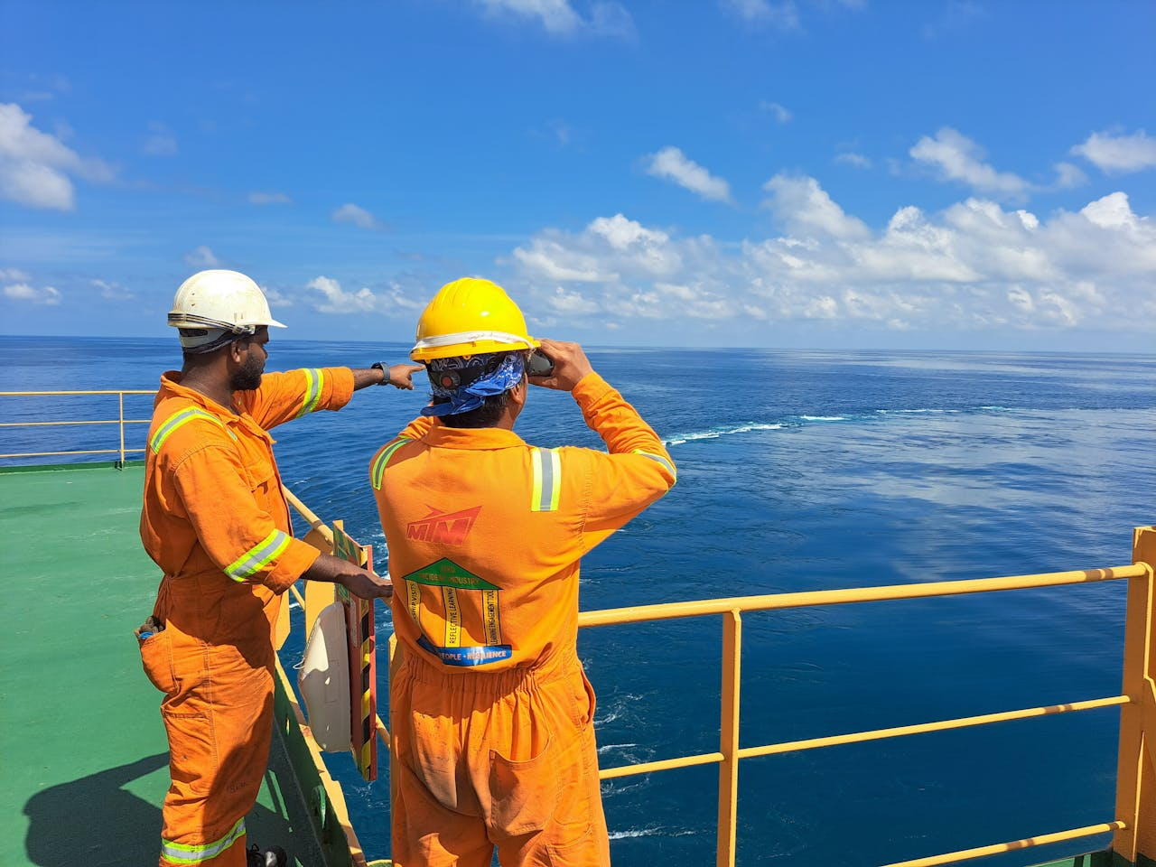 Home Two workers on an oil rig deck wearing safety gear inspect the ocean horizon under a clear sky.